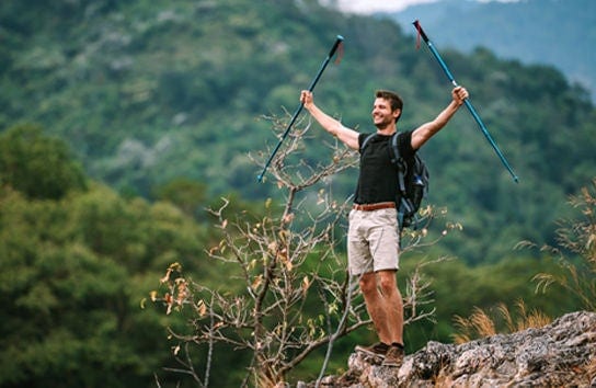 Hombre feliz con mochila y bastones de senderismo celebra en la cima de una montaña rocosa