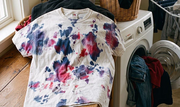 Playera blanca con manchas de tinte azul y rojo por desteñido junto a la lavadora en el cuarto de lavado.
