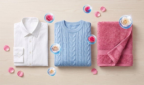 Ropa recién lavada y doblada en una cama de recámara, rodeada de burbujas flotantes con rosas que indican aroma fresco.