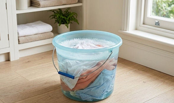 Cubeta transparente con agua jabonosa y ropa en remojo sobre el piso de madera del cuarto de lavado.